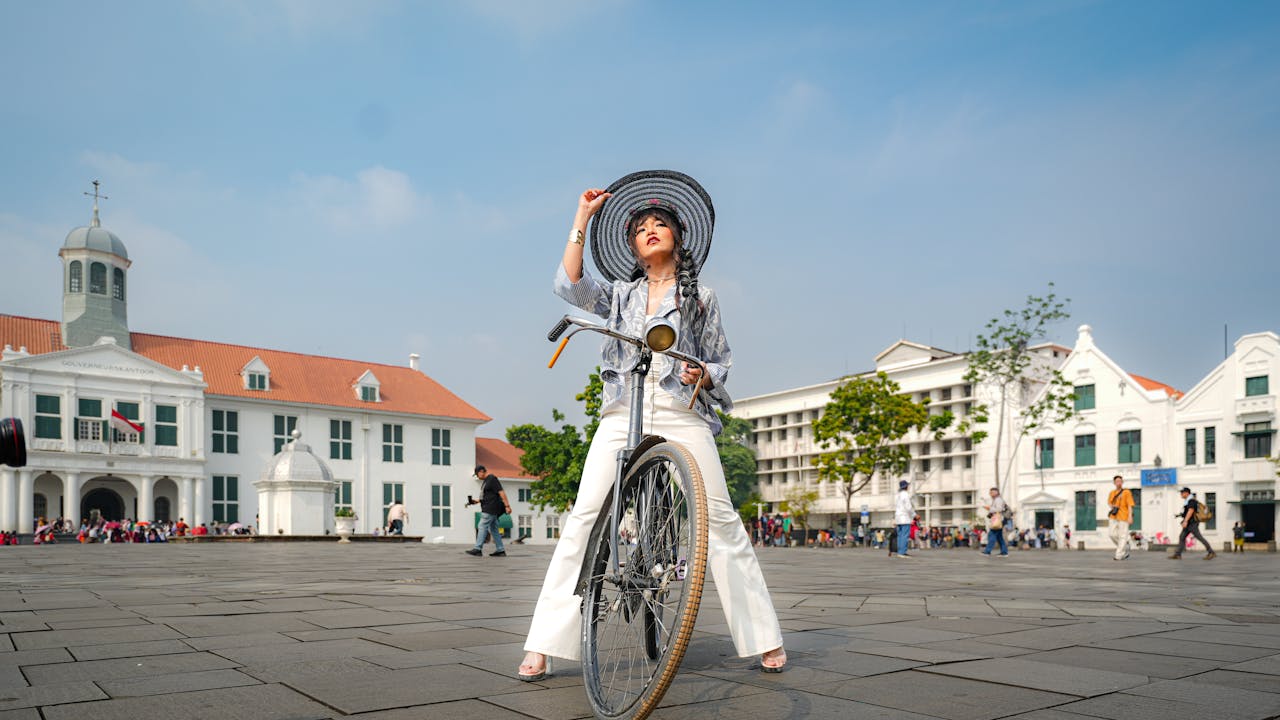 A fashionable woman poses with a vintage bicycle in Jakarta's Kota Tua, surrounded by historic colonial architecture.